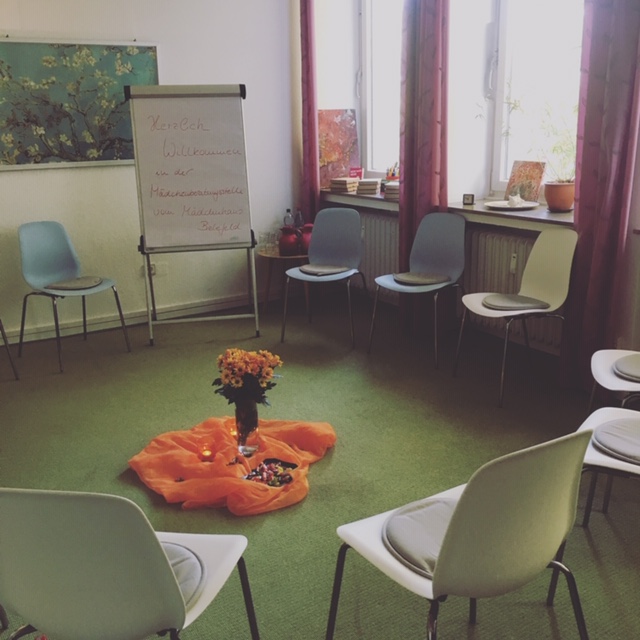 Auf dem Bild dsieht man einen Ausschnitt von einem Zimmer mit grünem Boden. Es sind Stühle reihum augstellt und in der Mitte liegt ein orangenes Tuch mit einem Strauß Blumen. Hinten an der Wand steht ein Flipchart.