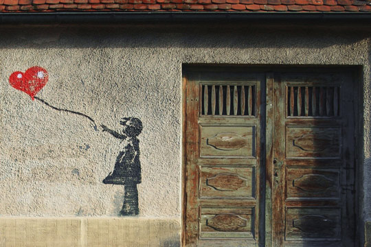 Das Foto zeigt eine graue Hauswand. Rechts im Bild ist eine braune Eichentür. Links daneben ist ein Graffiti, das ein Mädchen zeigt, dass einen Luftballon an einer Schnur in der Hand hält und diesem hinterher schaut.
