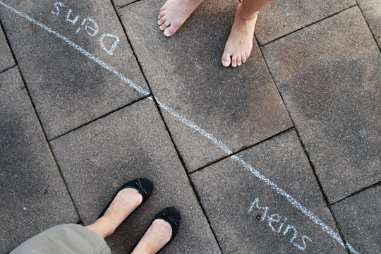 Das Foto zeigt Füße, die sich gegenüber vor einer Linie stehen. Auf der einen Seite steht 'Meins', auf der anderen 'Deins'.